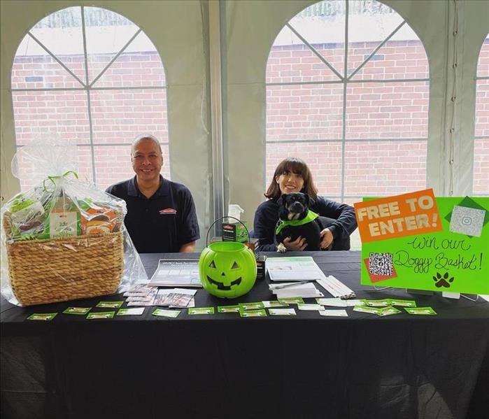 Two SERVPRO employees and a dog sit behind a black booth under a white tent.